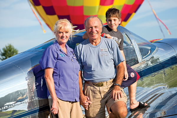 family of 3 standing in front of plane with a hot air balloon in the background
